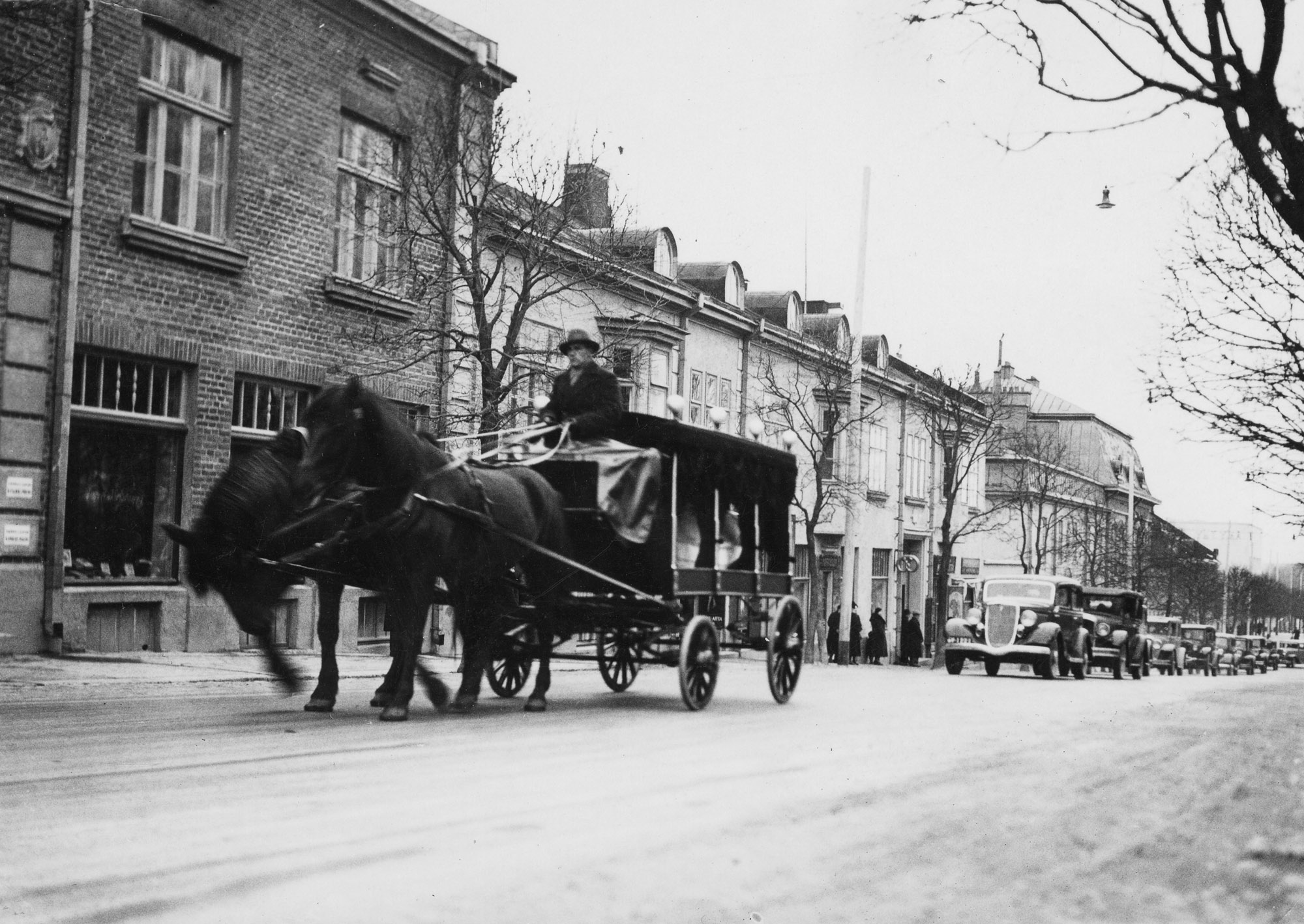 Mustavalkoinen valokuva hevosista vetämässä ruumisvaunuja.